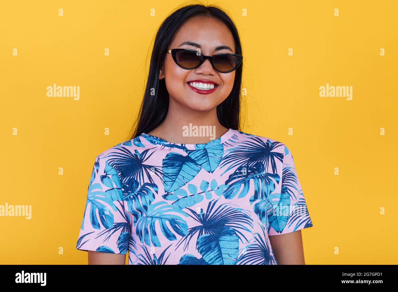 Donna asiatica felice in eleganti occhiali da sole e t-shirt con stampa a foglia tropicale guardando la fotocamera su sfondo giallo in studio Foto Stock