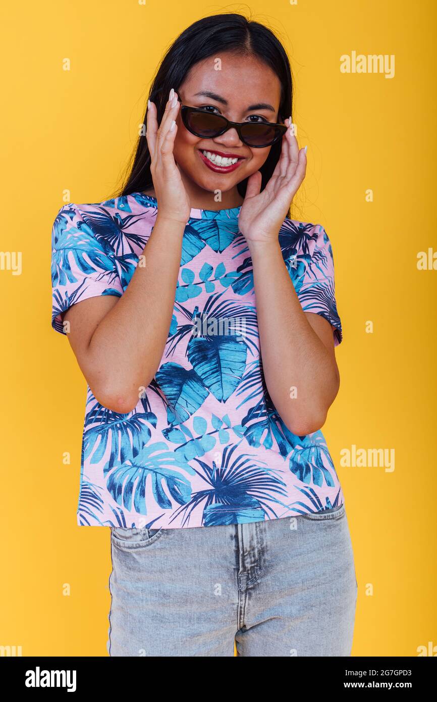 Donna asiatica felice in eleganti occhiali da sole e t-shirt con stampa a foglia tropicale guardando la fotocamera su sfondo giallo in studio Foto Stock