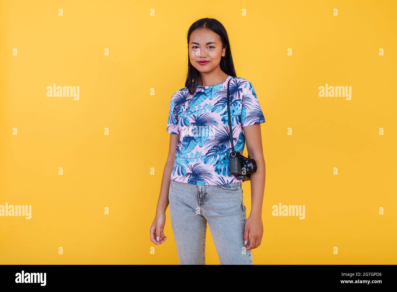 Donna asiatica felice in t shirt con stampa tropicale foglia con fotocamera su sfondo giallo in studio Foto Stock