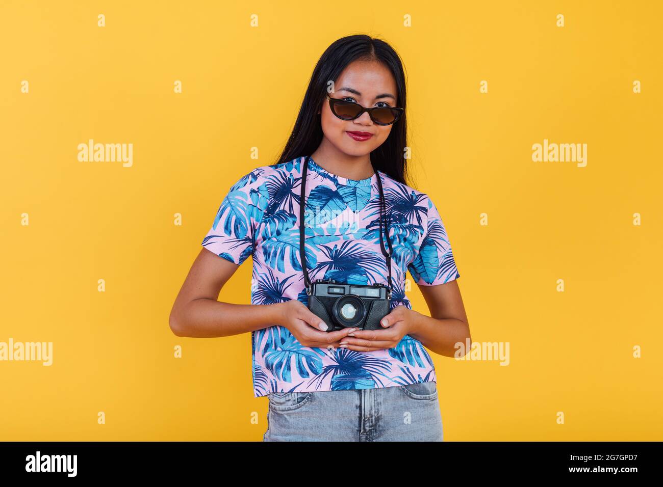 Donna asiatica felice in t shirt con stampa tropicale foglia che tiene fotocamera su sfondo giallo in studio Foto Stock