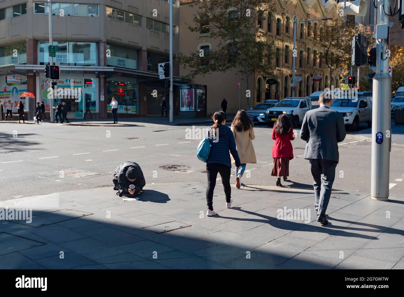 Le persone che attraversano un incrocio di strada nel centro di Sydney, Australia, camminano davanti a una persona inginocchiata e che tiene una tazza che inizia il giorno d'inverno Foto Stock