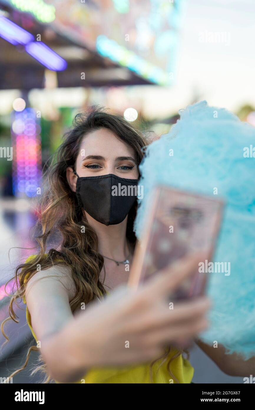 Donna in maschera protettiva e con caramelle di cotone dolce blu che si autosshot sul telefono cellulare mentre si divertono al campo da golf Foto Stock