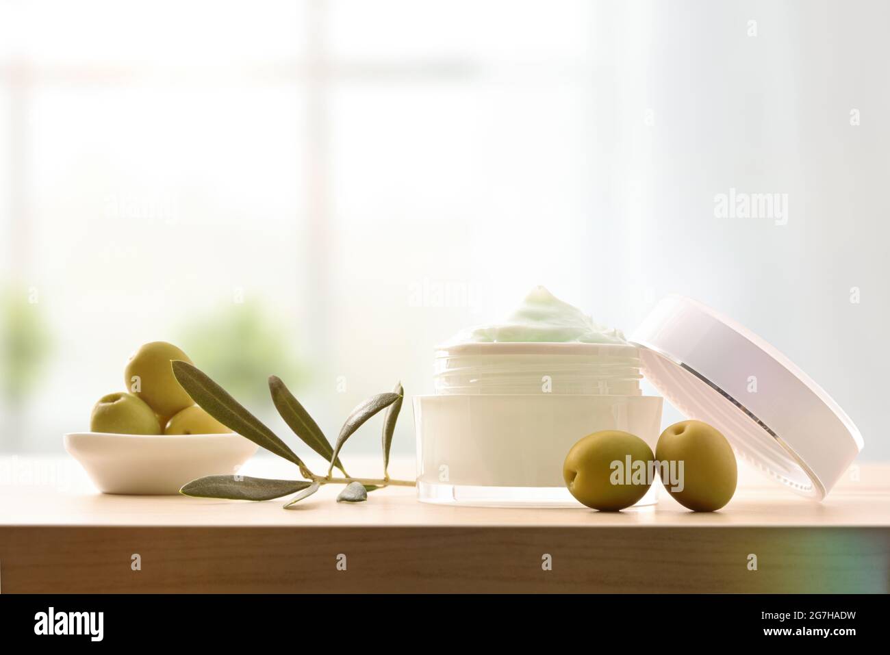 Crema cosmetica idratante con estratto di oliva per la pelle con rametto e olive su tavola di legno in camera. Vista frontale. Foto Stock