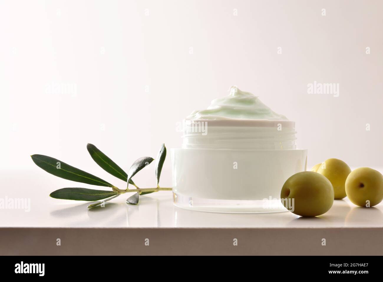 Crema cosmetica idratante con estratto di oliva per la pelle con rametto e olive su tavola bianca. Vista frontale. Foto Stock
