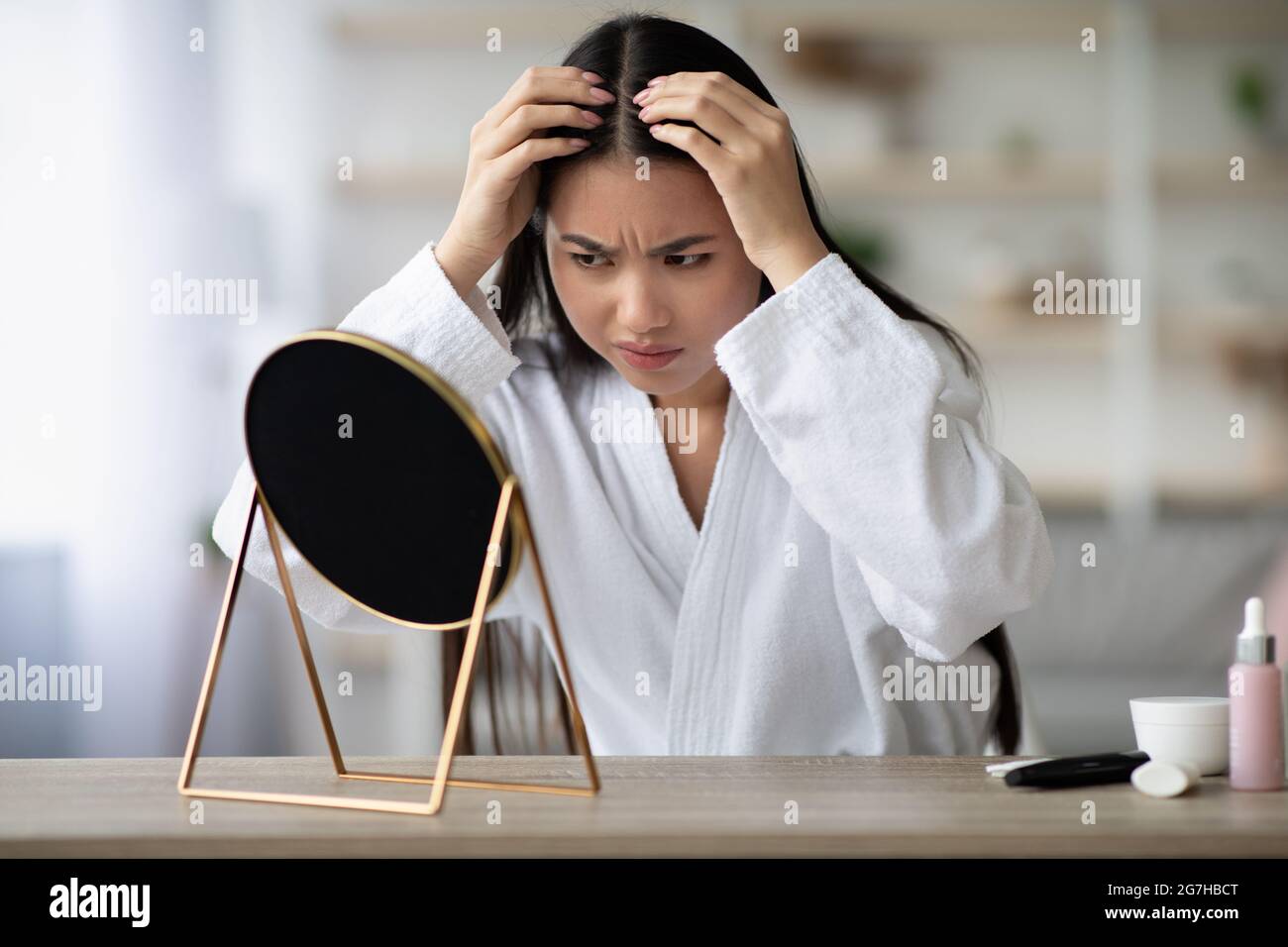 Arrabbiato giovane donna asiatica in accappatoio bianco seduta in camera da letto, controllando i suoi capelli grigi, guardando lo specchio, dopo aver deluso espressione del volto, copia spac Foto Stock