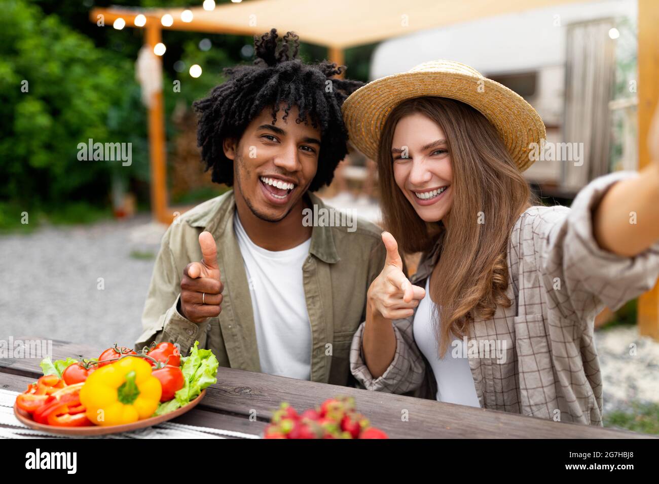 Giovane coppia multinazionale con pranzo insieme al campeggio, in posa per selfie vicino camper van, puntando alla macchina fotografica e sorridendo. Ragazzo nero freddo e hi Foto Stock