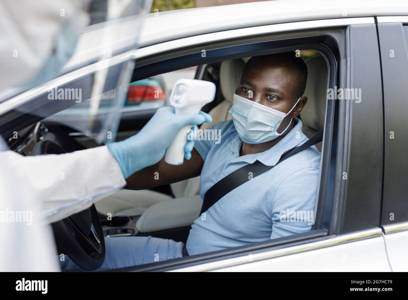 Uomo nero driver indossare maschera protettiva viso, seduto all'interno di auto bianca, lavoratore medico in tuta di protezione controllare il temperamento corpo di ragazzo afroamericano Foto Stock