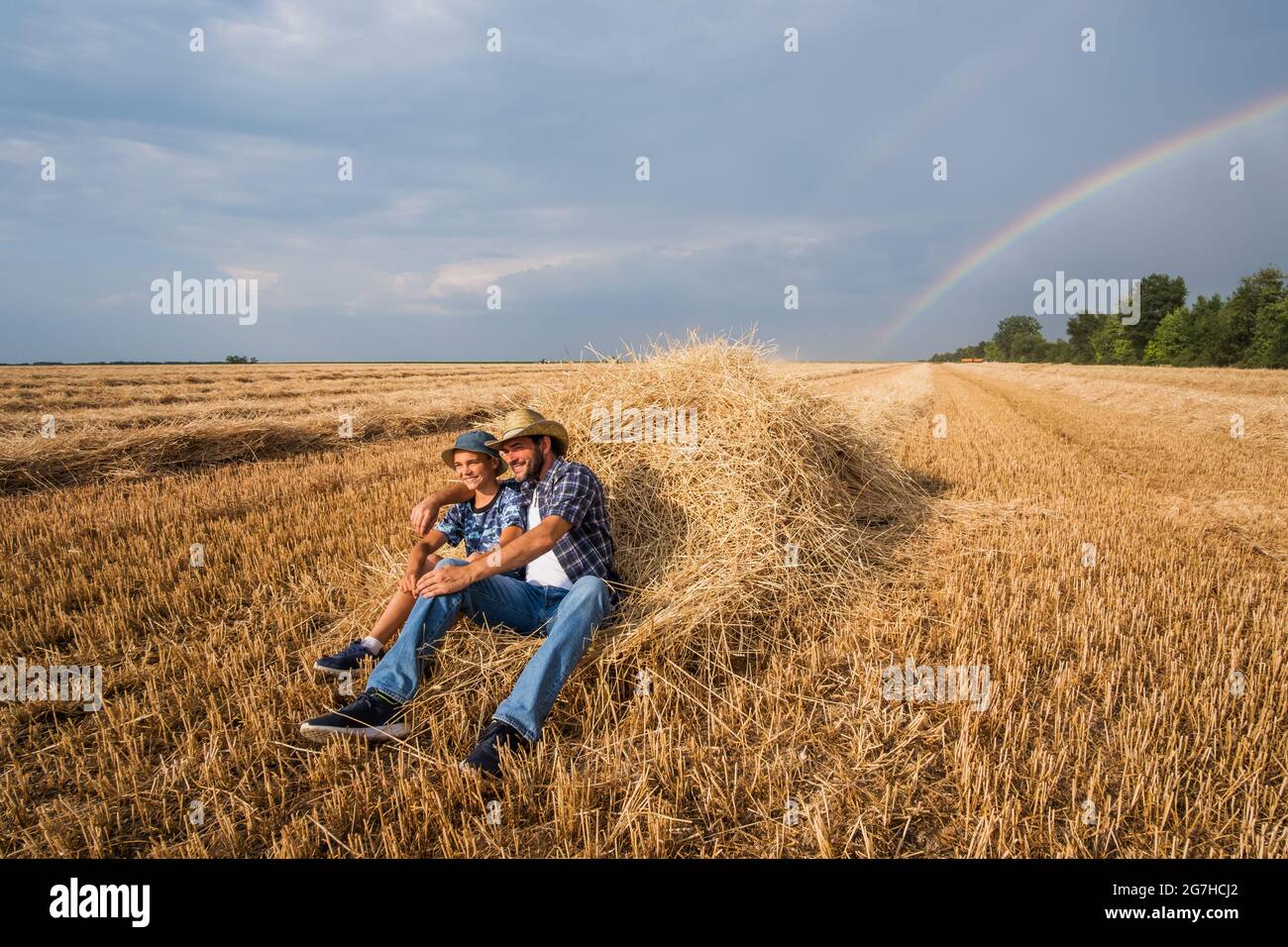 Padre e figlio riposano dopo il raccolto riuscito. Arcobaleno nel cielo dietro di loro. Foto Stock
