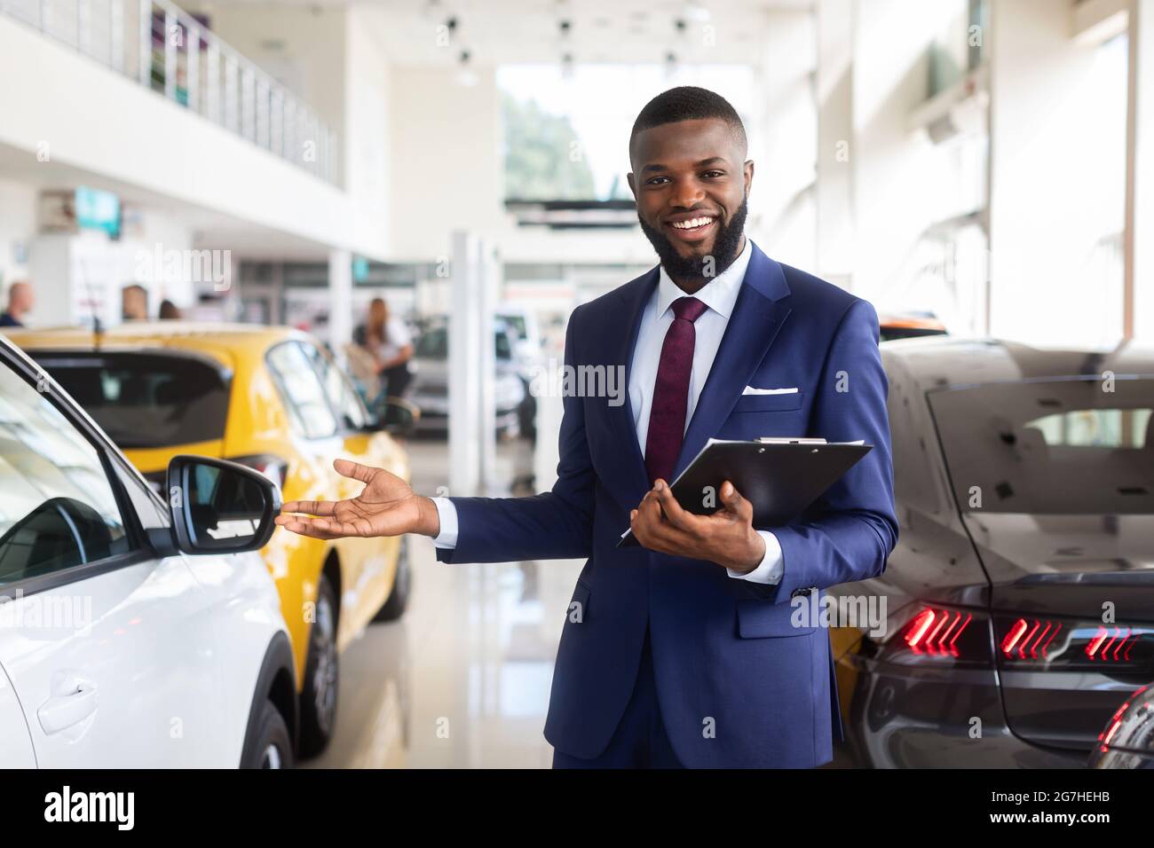 Ritratto del bel venditore afroamericano al posto di lavoro in Car Showroom, felice sorridente Black Auto Dealership Manager in suit con Clipboard a Han Foto Stock