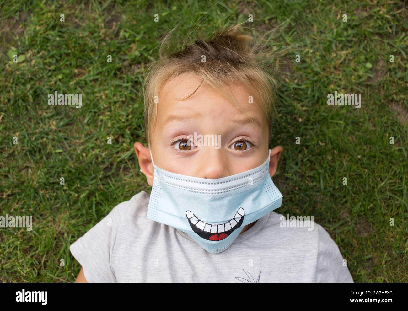 il bambino in una maschera medica blu con un sorriso dipinto su di esso giace sull'erba con ampi occhi aperti. Atteggiamento positivo durante la pandemia di COVID 19. W Foto Stock