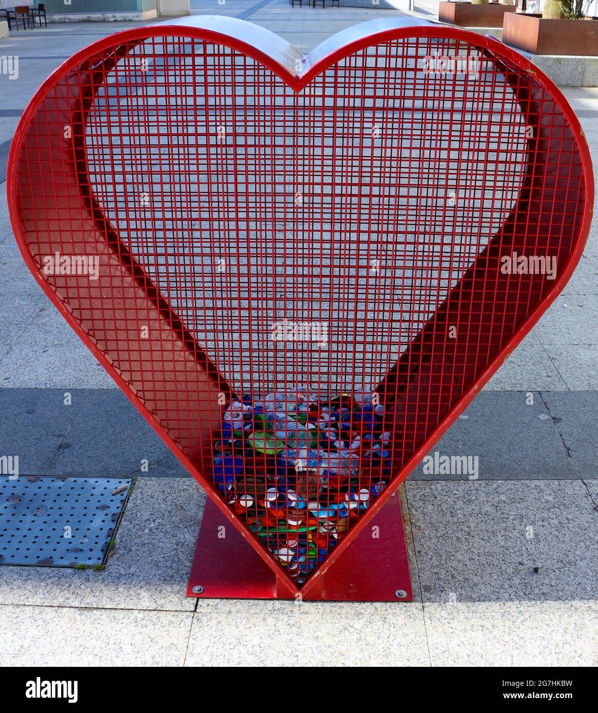 Contenitore rosso a forma di cuore per il riciclaggio di tappi in plastica in una strada a Santona Cantabria Spagna Foto Stock