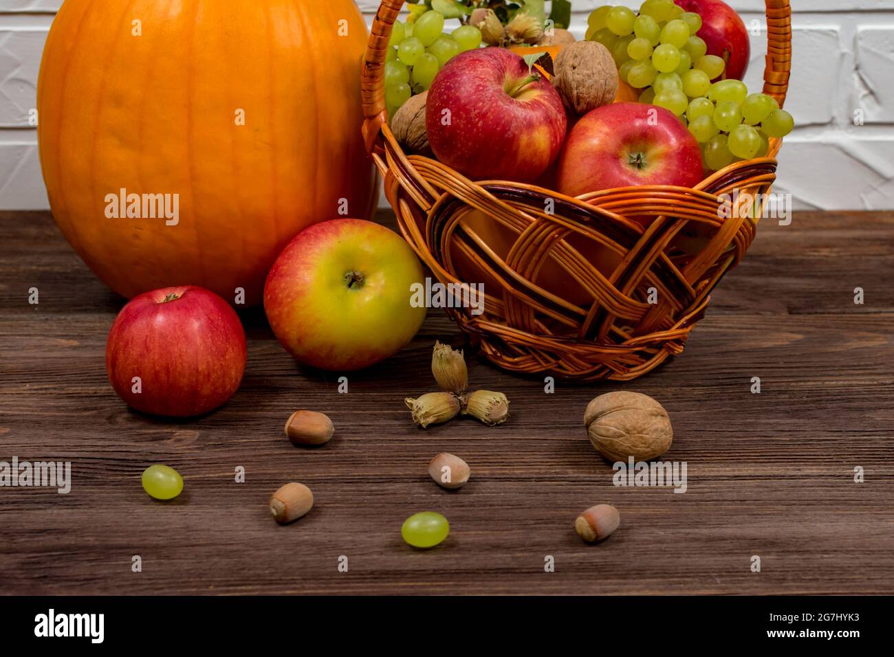 Zucche d'arancia con mele e pere in un cesto di vimini con uva, noci e foglie su un tavolo di legno marrone contro un muro di mattoni bianchi Foto Stock