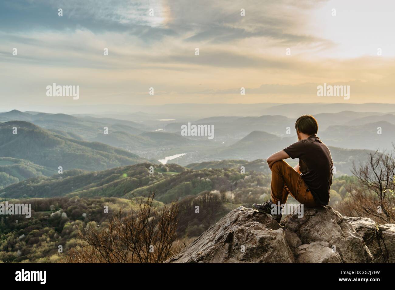 Il viaggiatore maschile con la mente tranquilla seduto sulla roccia godendo di vedute della lussureggiante valle di primavera al tramonto. Giorno di escursioni lifestyle attivo.Wanderlust donna rilassante Foto Stock