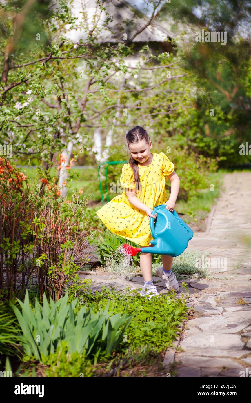 una ragazza sorridente e gioiosa in un vestito giallo che vola nel vento sta innaffiando i fiori giovani da una lattina di irrigazione blu nel cortile anteriore di una casa in un Foto Stock