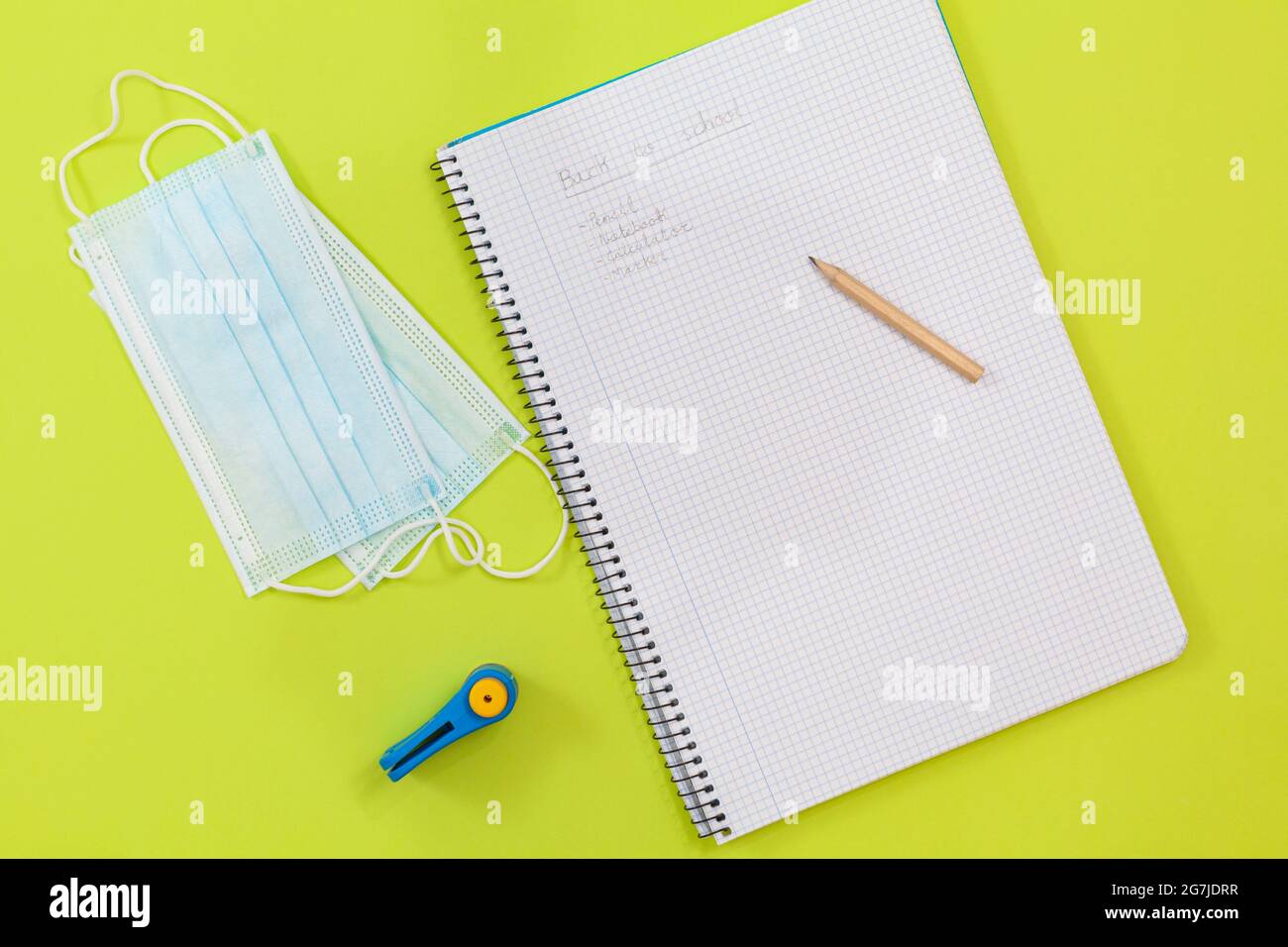 Su uno sfondo verde chiaro sono due maschere chirurgiche, un punzone e un quaderno con una serie di materiali scolastici scritti in matita. Foto Stock