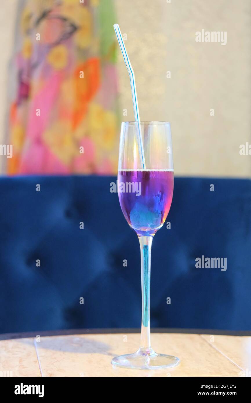un bicchiere di champagne in piedi su un tavolo con una paglia e una bevanda di una tonalità viola sullo sfondo di una sedia blu Foto Stock