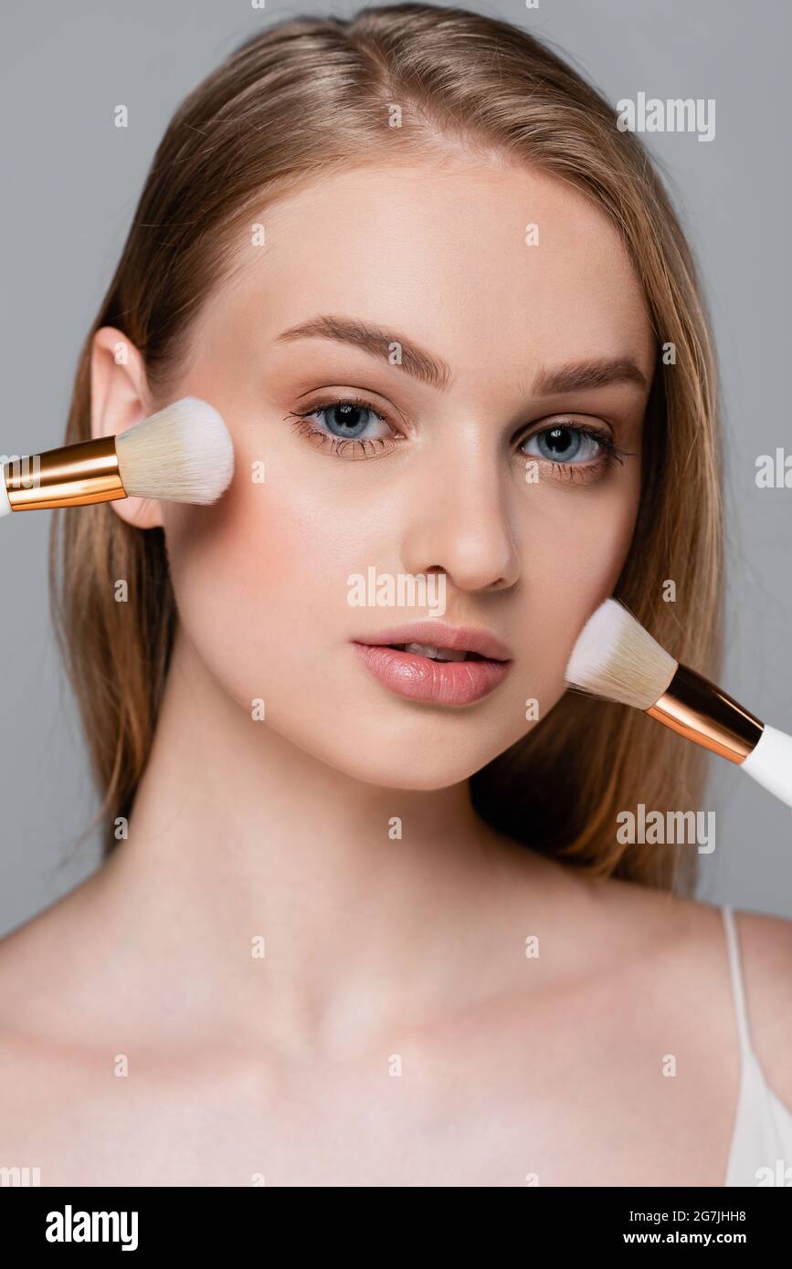 primo piano di giovane donna che tiene diverse spazzole cosmetiche e l'applicazione di polvere viso isolato su grigio Foto Stock