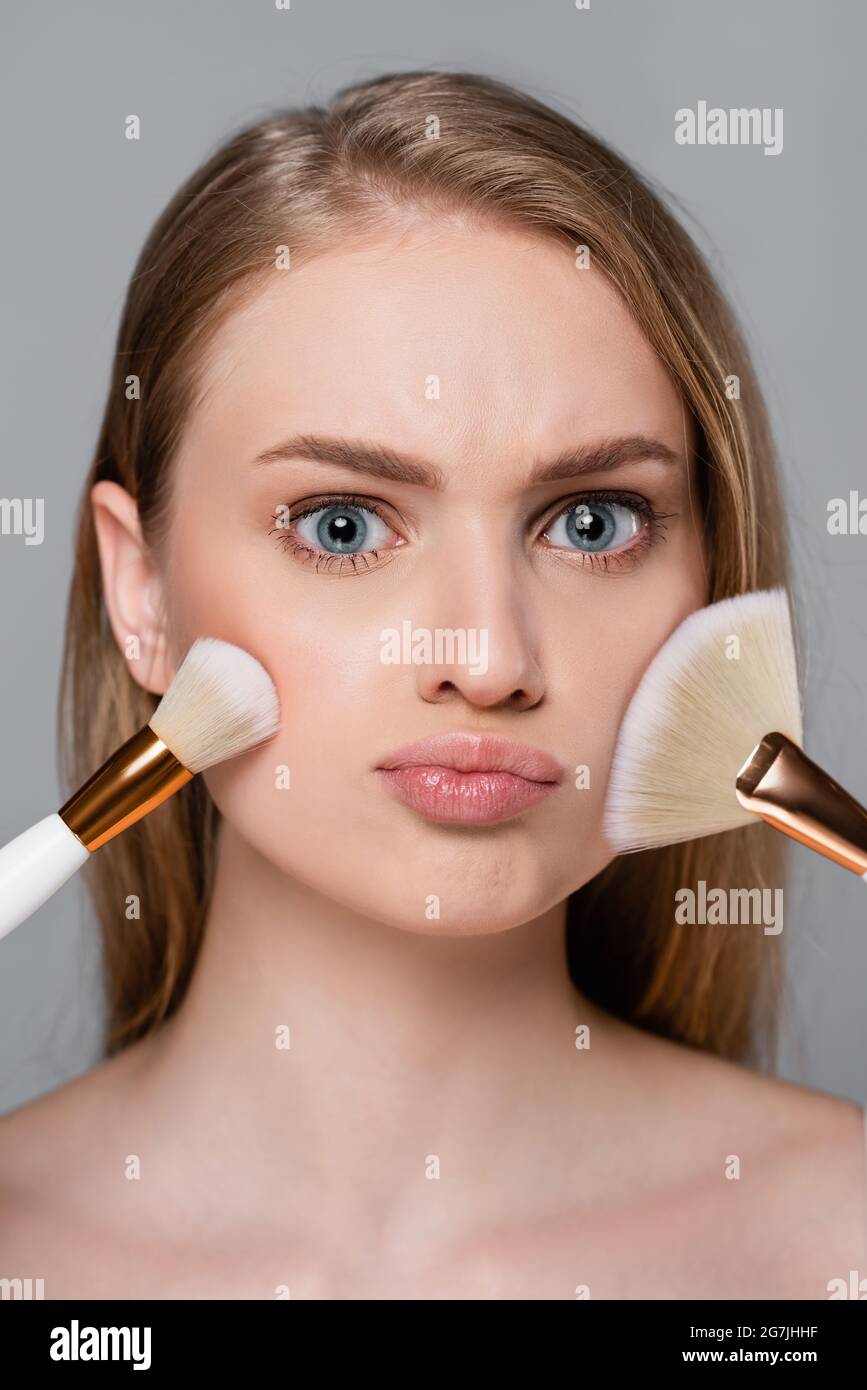 giovane donna che tiene le spazzole cosmetiche ed applica la polvere del viso isolata su grigio Foto Stock