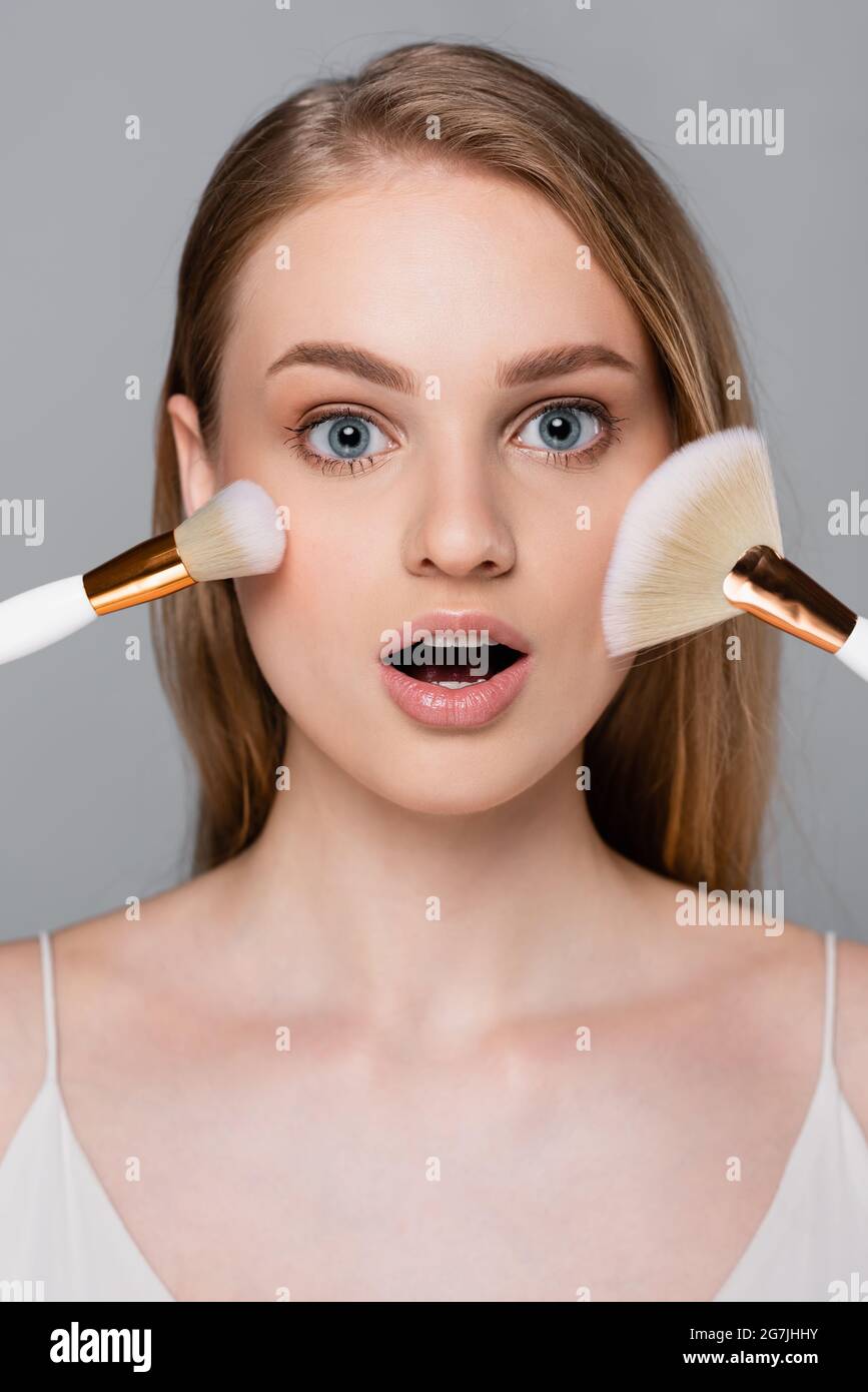 stupito giovane donna che tiene le spazzole cosmetiche vicino faccia isolata su grigio Foto Stock