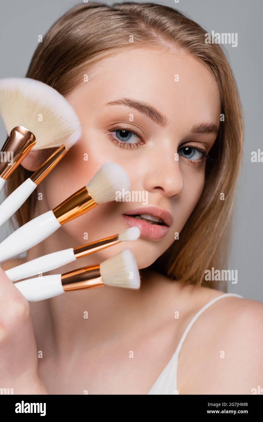 giovane donna che tiene set di spazzole cosmetiche diverse mentre si guarda la fotocamera isolata su grigio Foto Stock