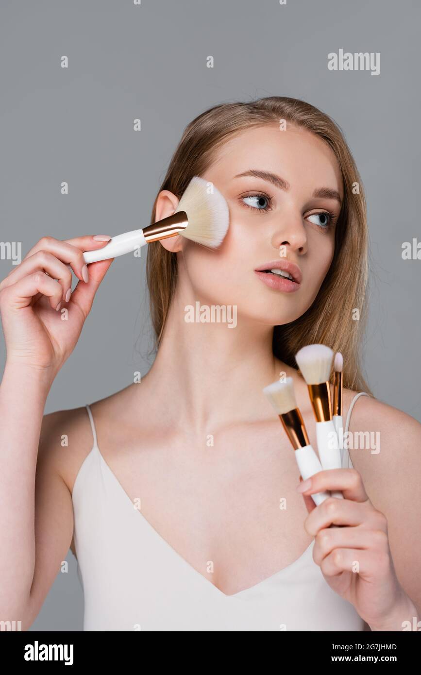 giovane donna che tiene insieme di spazzole cosmetiche diverse e l'applicazione di polvere viso isolato su grigio Foto Stock