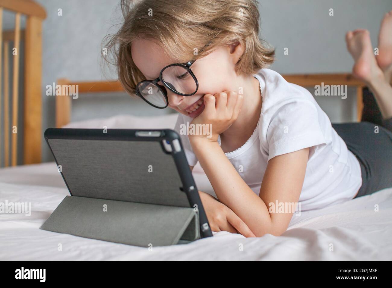 La bambina con gli occhiali fa i compiti in linea, sdraiata sul letto a casa. Comunicazione con i parenti online. Il concetto dell'inizio del nuovo Foto Stock