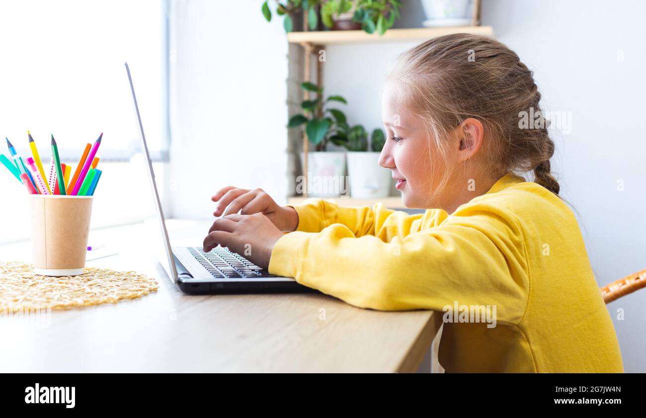 Caucasica bionda scuola ragazza, digitando su una tastiera portatile mentre si siede vicino alla finestra a casa, blogging, chattare, visualizzare le pagine dei social media o. Foto Stock