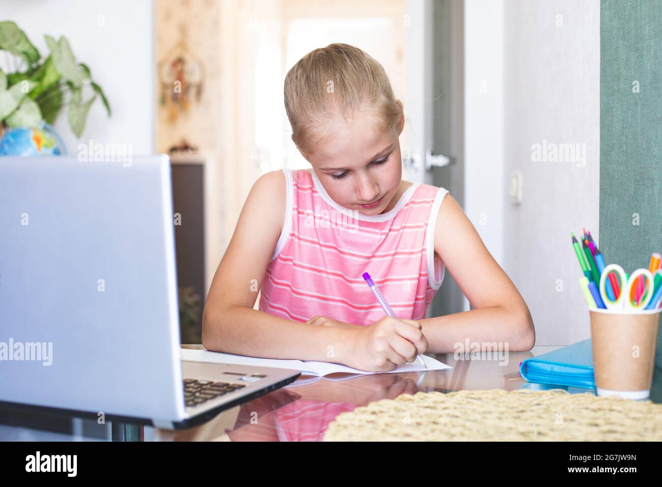 Ragazza caucasica di 10-11 anni facendo compiti vicino a un computer portatile, seduto alla scrivania scrive in un notebook. Apprendimento a casa. Ritorno a scuola. Accogliente Foto Stock
