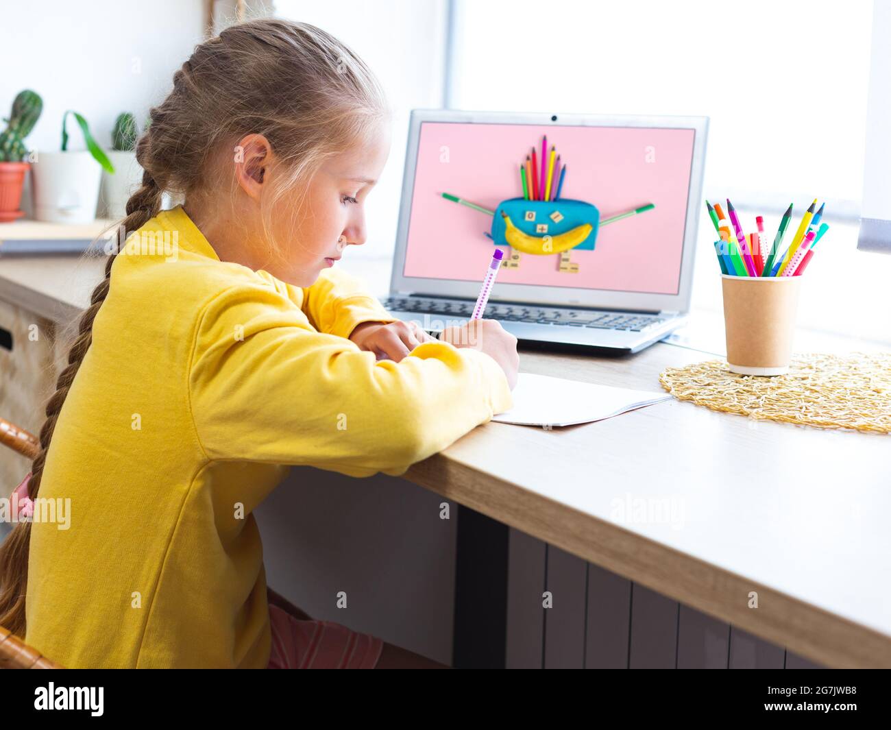 La studentessa caucasica fa i compiti, disegna in un taccuino. Posto di lavoro accogliente vicino alla finestra. Forniture scolastiche multicolore, forniture per ufficio e una banana in Foto Stock