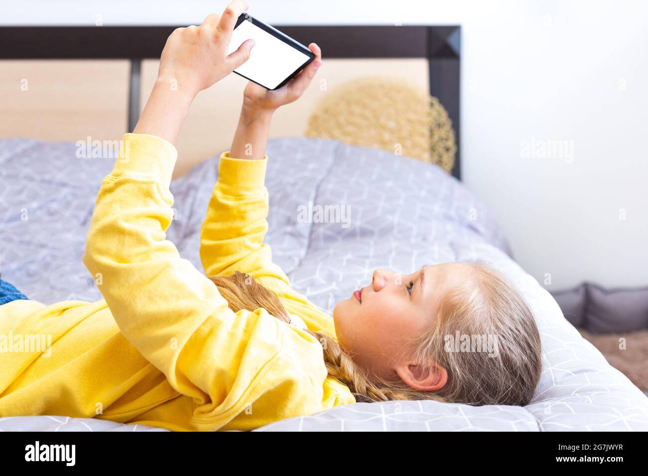 Ragazza caucasica adolescente in una felpa gialla, sdraiata sul letto con uno smartphone in mano, giocare, shopping online, chattare, blogging Foto Stock