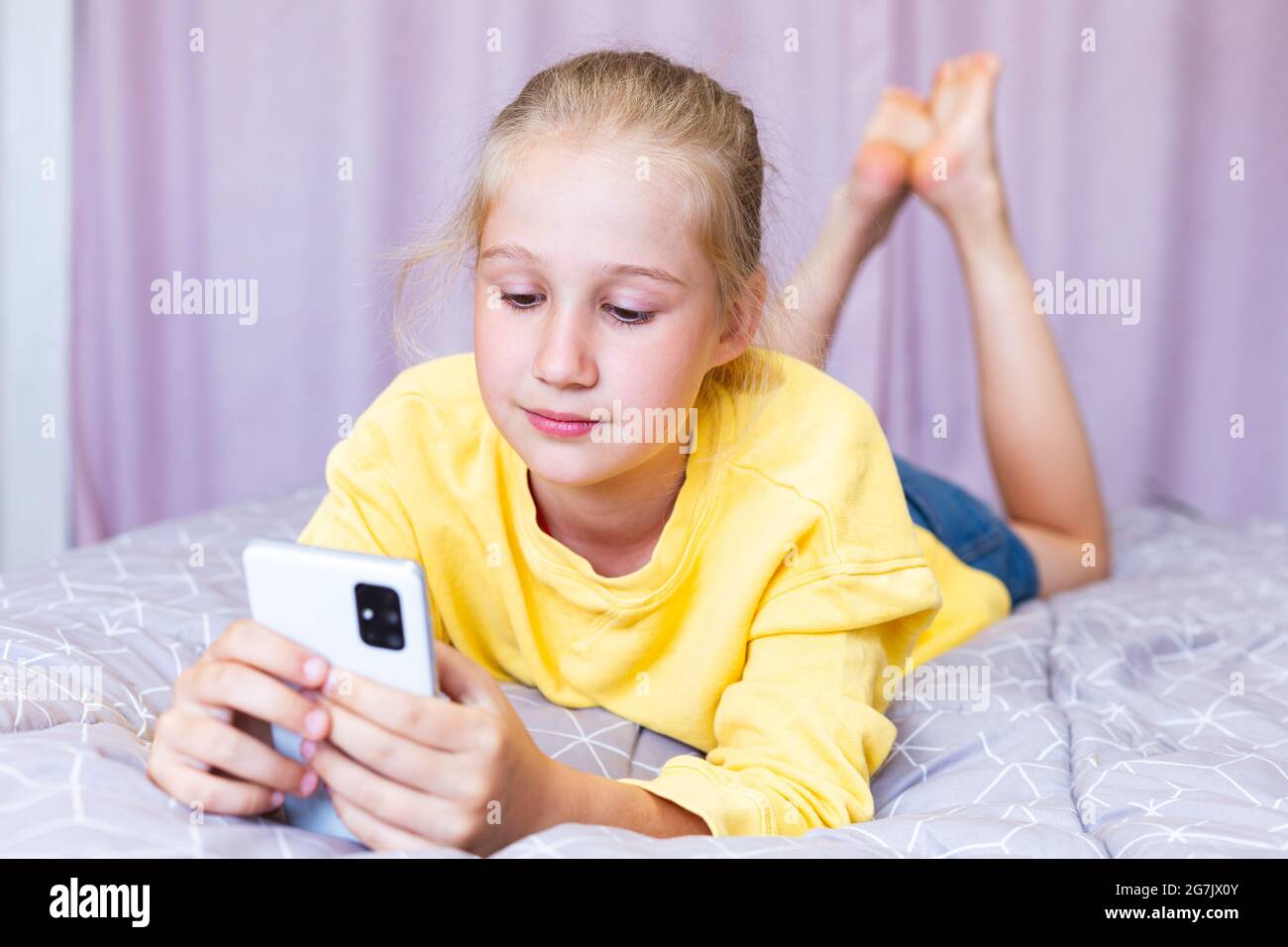Una ragazza caucasica bionda adolescente si trova sul letto con un telefono nelle sue mani, gioca giochi, shopping online, blogging. Comunicazione sui social network Foto Stock