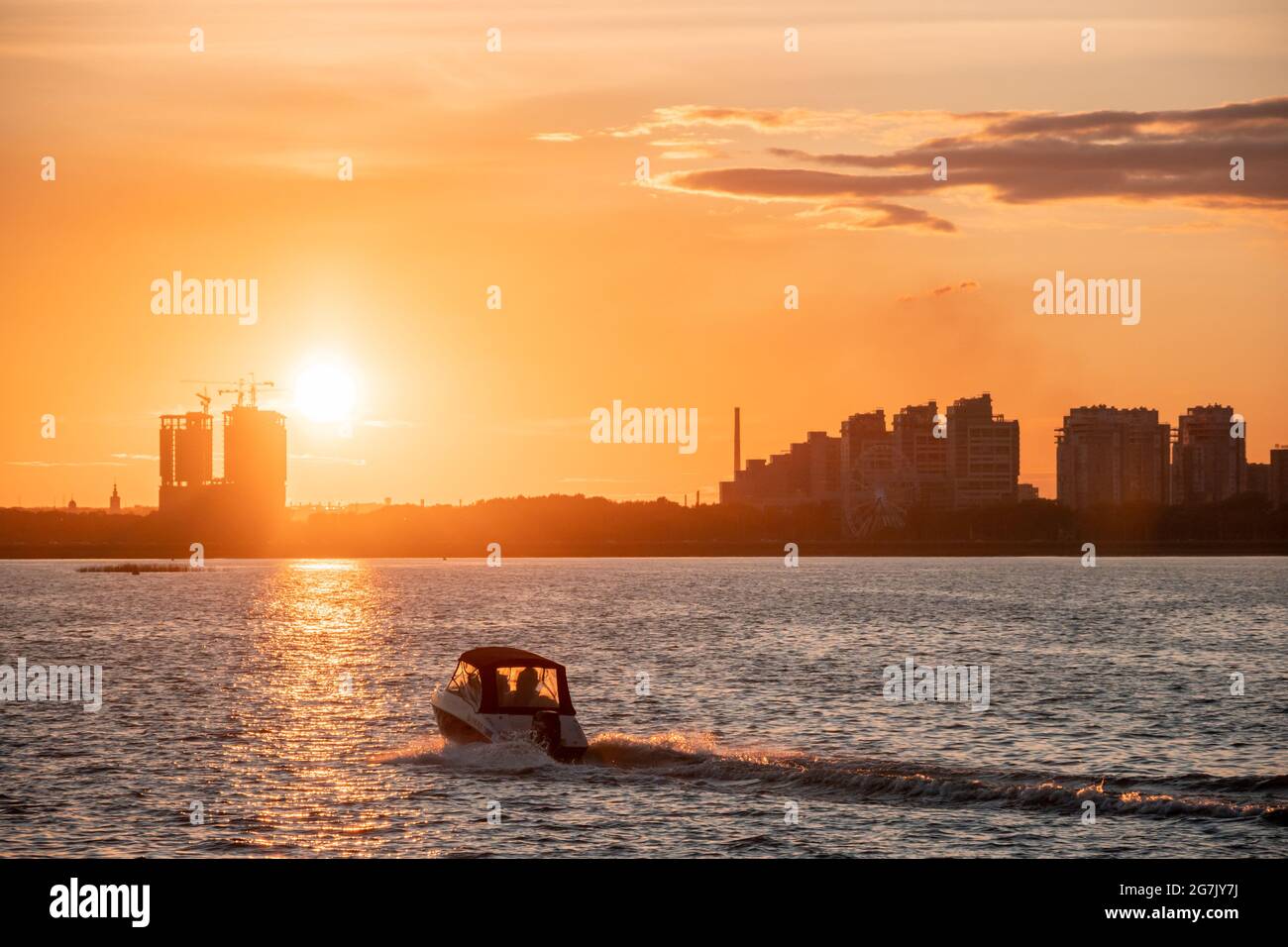 Silhouette di edifici cittadini sulla riva sullo sfondo del sole che tramonta e baglia l'acqua. Tramonto sul fiume, una barca a motore crossin Foto Stock