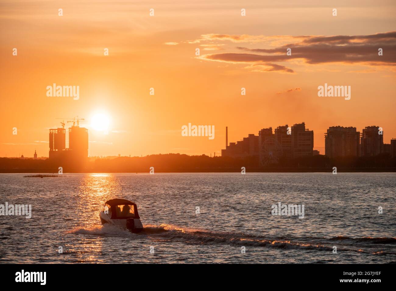 Silhouette di edifici cittadini sulla riva sullo sfondo del sole che tramonta e baglia l'acqua. Tramonto sul fiume, una barca a motore crossin Foto Stock