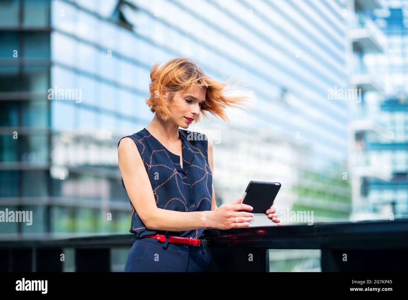 Attraente donna elegante in piedi guardando i media o leggendo online sul suo tablet all'aperto su un ponte urbano con sfondo grattacielo e capelli blowi Foto Stock