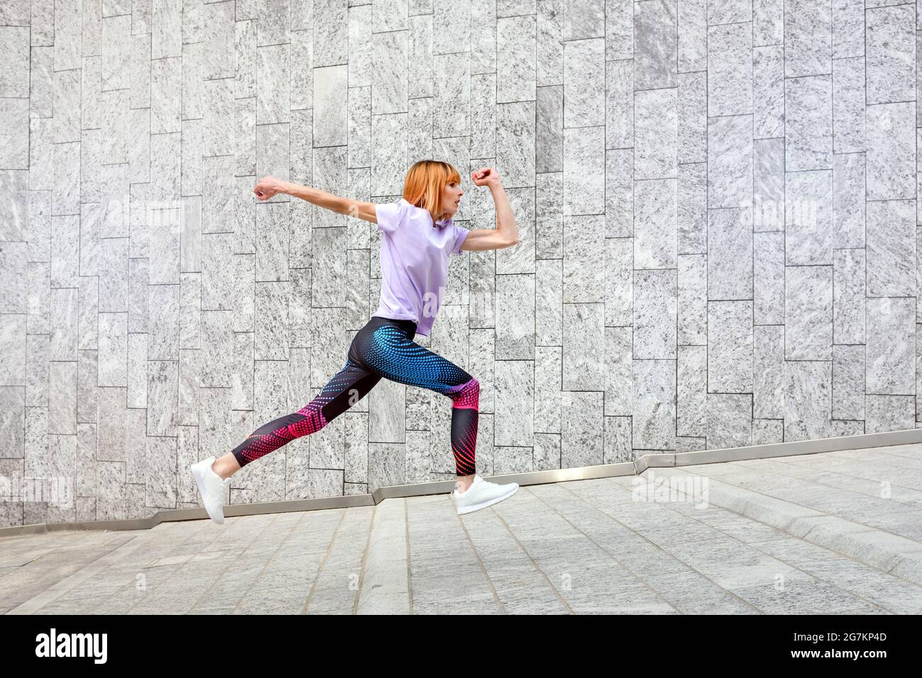 Donna sportiva matura che fa jogging attraverso la città correndo su un pendio passato edifici commerciali in un sano stile di vita attivo all'aperto e concetto di fitness Foto Stock