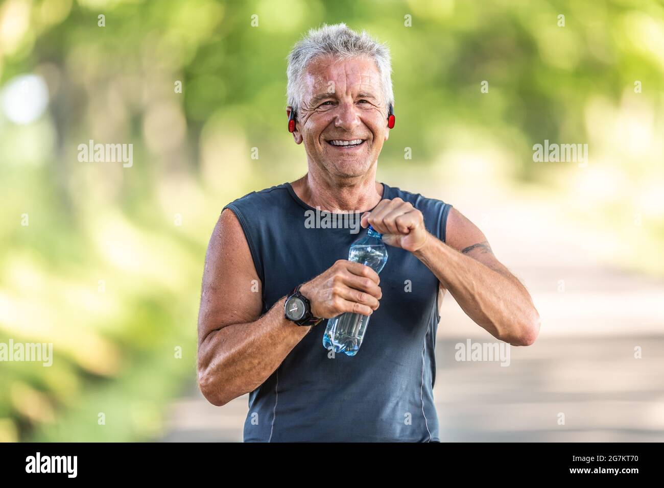 Un uomo sorridente, in forma e in pensione con i capelli grigi sorride mentre apre una bottiglia d'acqua dopo aver terminato un allenamento aerobico. Foto Stock