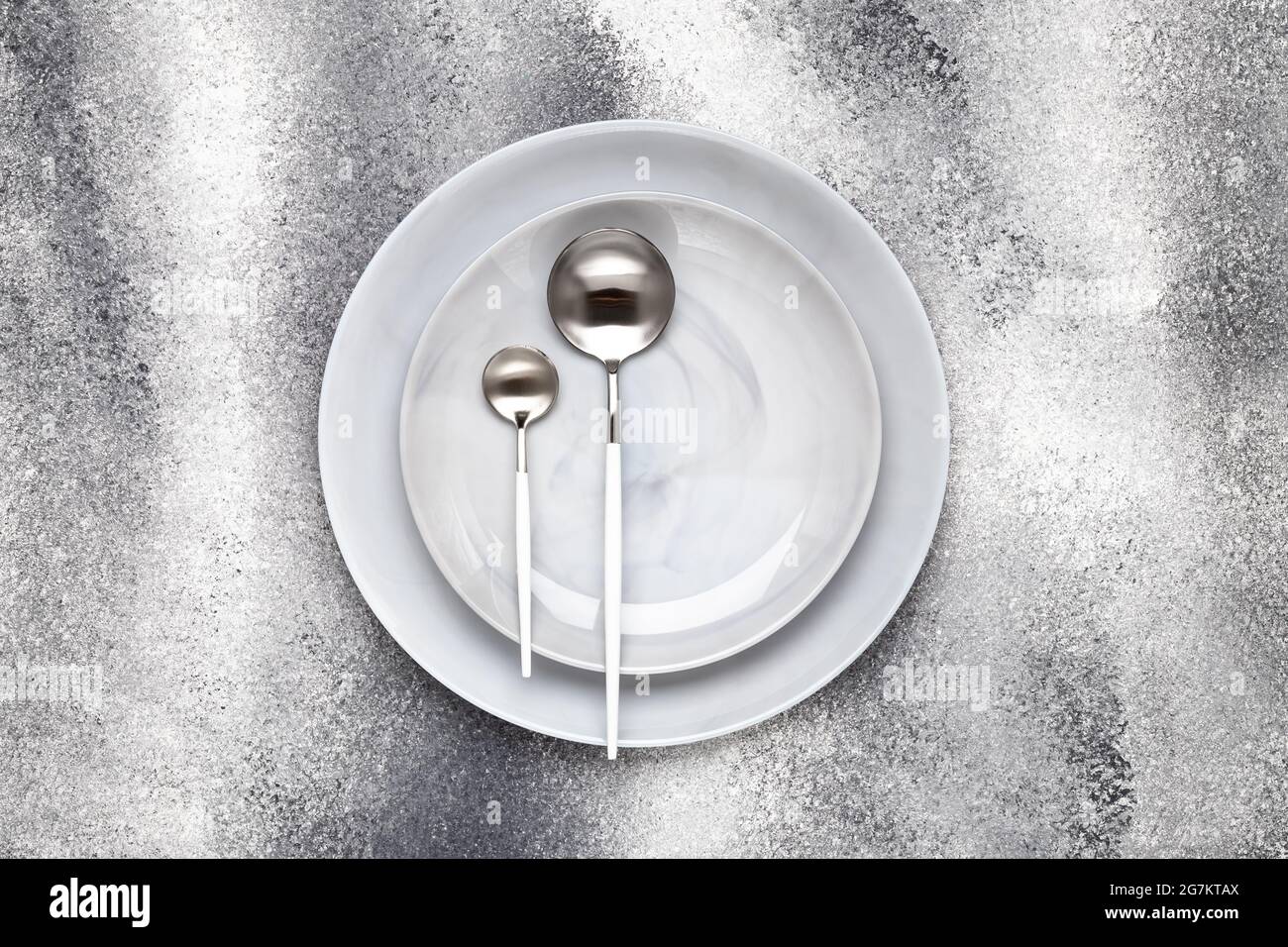 Piastra vuota in ceramica e cucchiaino da tè inutilizzato con cucchiaio da tavola, posate su un tavolo grigio, concetto di servizio. Design piatto. Menu del ristorante. Biglietto d'auguri, l Foto Stock