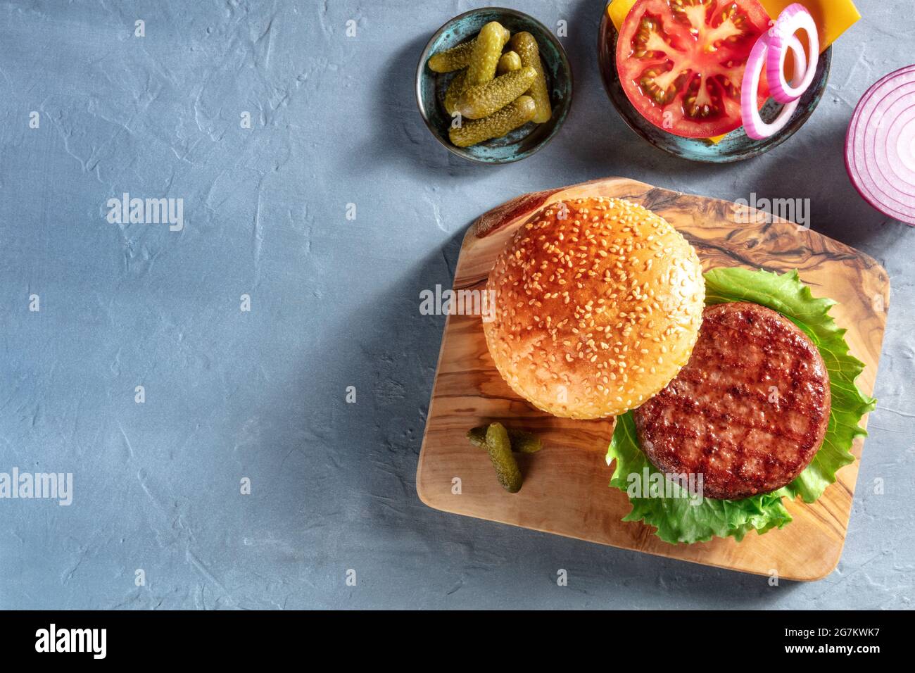 Ingredienti Burger. Hamburger paty, sesamo panino, m pomodoro, cipolla e sottaceti, sparato dall'alto su uno sfondo blu con spazio copia. Ricetta di hamburger Foto Stock