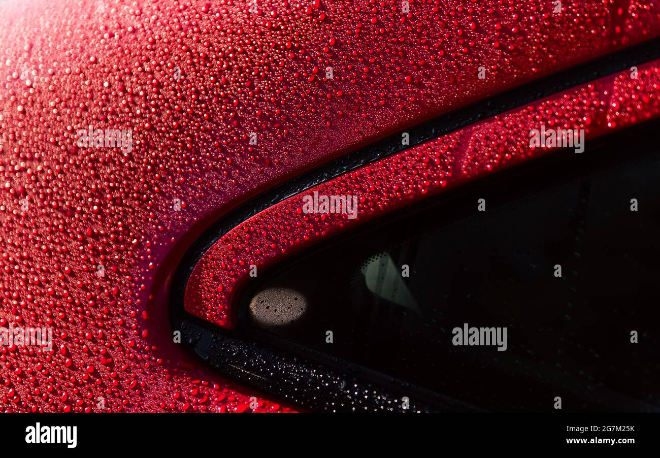 Gocciolina d'acqua sull'auto. Bagne d'acqua dopo la pioggia o il lavaggio dell'auto su una superficie di vernice lucida rossa. Sfondo goccia d'acqua. L'acqua cade macro su una superficie rossa. Foto Stock