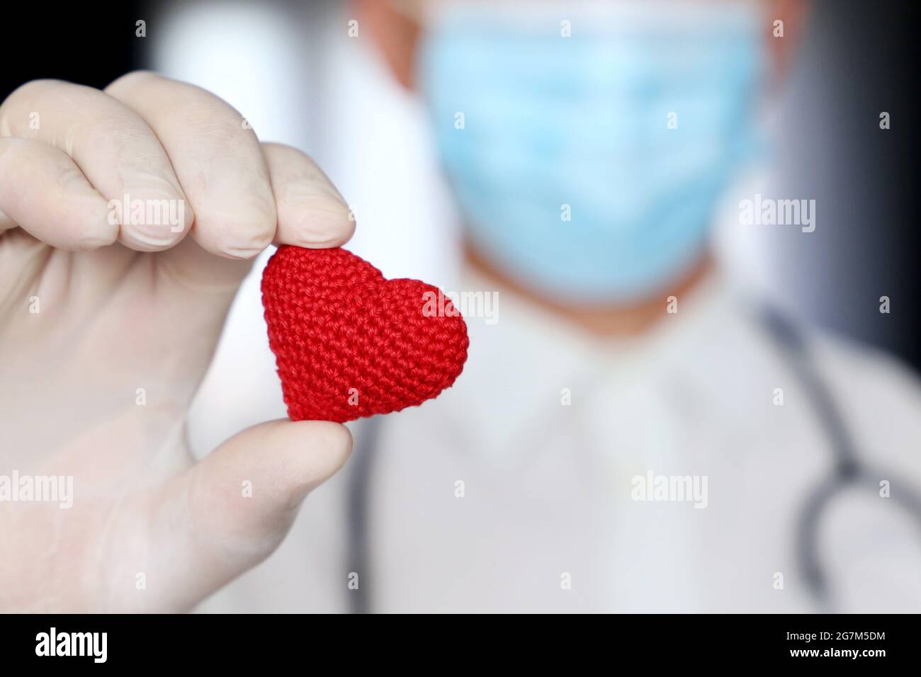 Cuore lavorato a maglia rosso in mano di medico uomo in maschera medica. Concetto di assistenza sanitaria, cardiologia, salvataggio di vite umane durante una pandemia di coronavirus covid-19 Foto Stock