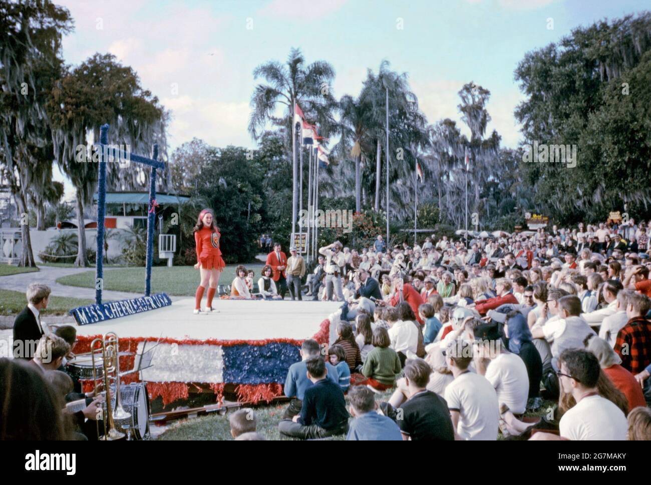 Una ragazza adolescente, vestita tutta in rosso, si esibisce in un palcoscenico all'aperto sotto il sole della sera in un concorso ‘MBiss Cheerleader USA’ a Cypress Gardens, Polk County, Florida, USA nel 1968. Sta cantando (o allegorando o cantando) accompagnato da una band (in basso a sinistra), con fotografi presenti davanti ad un pubblico di grandi dimensioni. Questa immagine proviene da un vecchio lucido a colori amatoriale americano Kodak – una fotografia d'epoca degli anni '60. Foto Stock