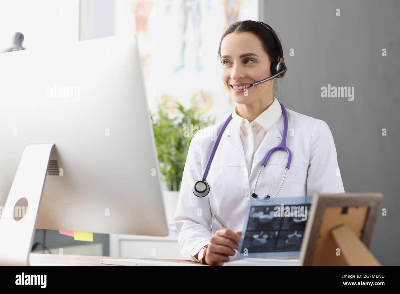 Medico in cuffie con microfono che tiene raggi X davanti allo schermo del computer Foto Stock