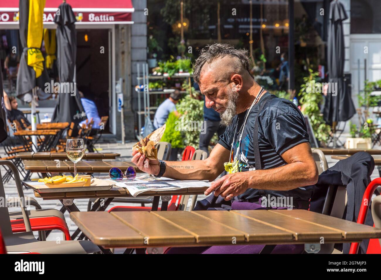 Vecchio punk-stile uomo leggere giornale e mangiare patatine al bar ristorante all'aperto tavolo a Saint Gilles, Bruxelles, Belgio. Foto Stock