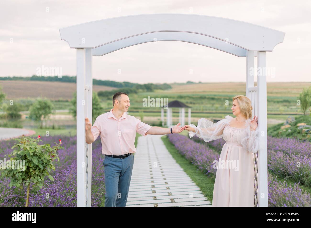 Felice insieme. Bella coppia elegante di mezza età nel campo di lavanda, in piedi e guardando l'un l'altro, appoggiandosi sul grande arco di legno bianco. Primo piano verticale Foto Stock