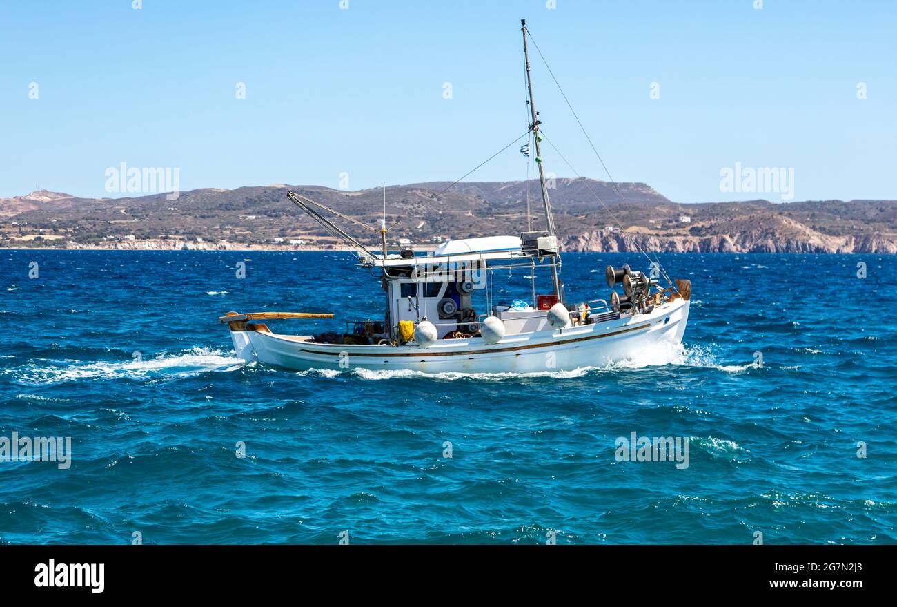 CICLADI, Grecia. Barca da pesca bianca tradizionale barca in legno vela in mare agitato. Terra rocciosa, costa dell'isola greca e cielo azzurro Foto Stock