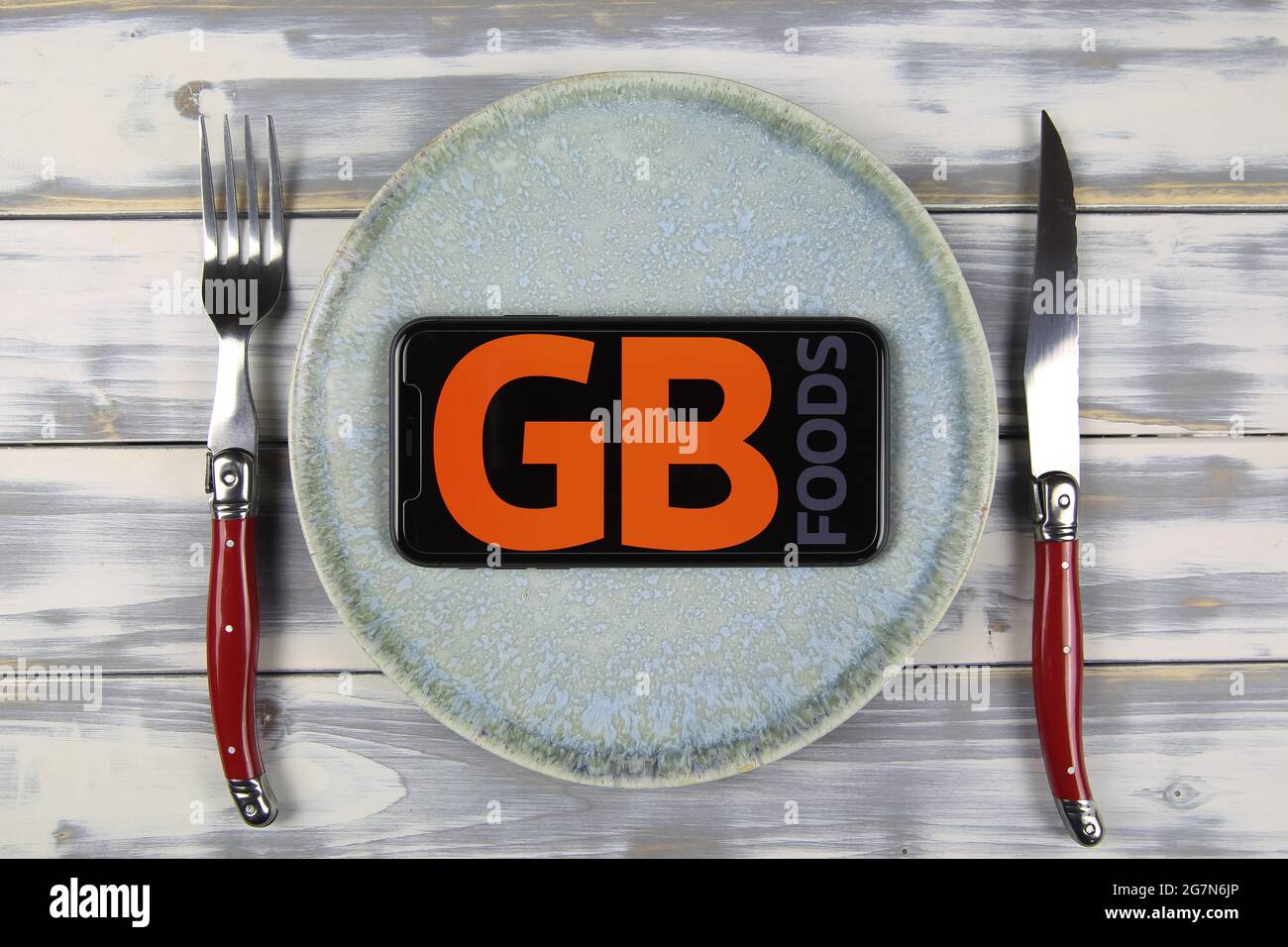 Viersen, Germania - 9 maggio. 2021: Closeup di telefono cellulare su piastra e tavolo in legno, posate con logo scritta della società spagnola gb alimenti (Focus su ce Foto Stock