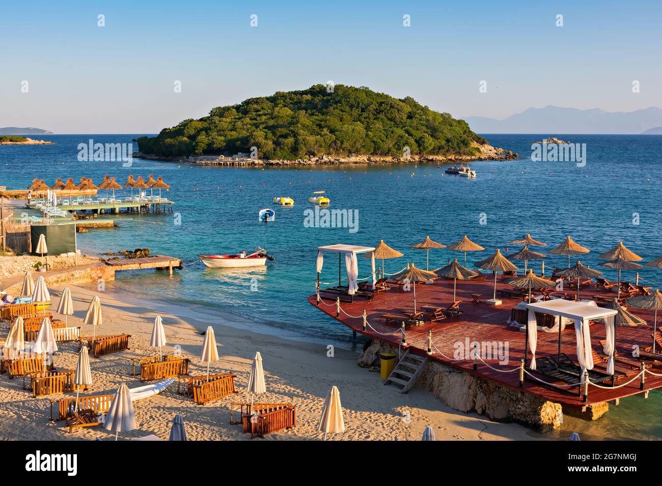 Splendido paesaggio mattutino con spiaggia di sabbia, isola, ombrelloni e blu Mar Ionio sulla costa albanese Foto Stock