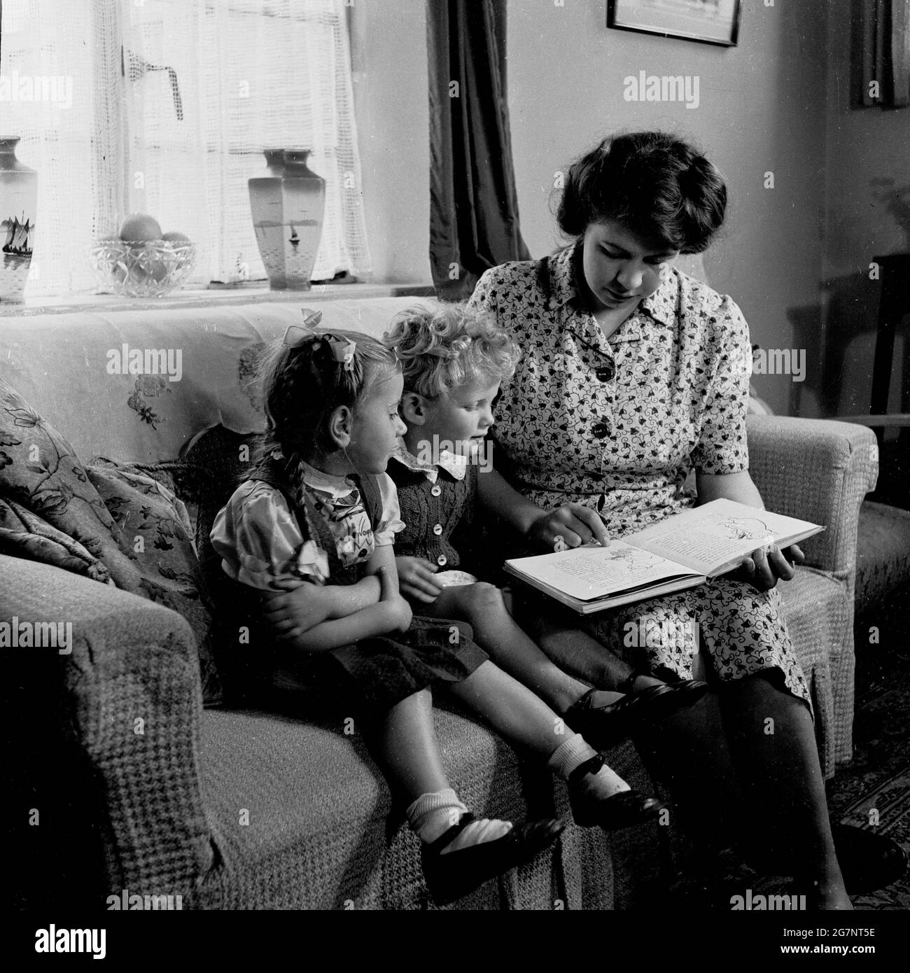 Anni '50, storico, una madre seduta su un divano con i suoi due bambini piccoli che li legge una storia da un libro, Inghilterra, Regno Unito. Foto Stock