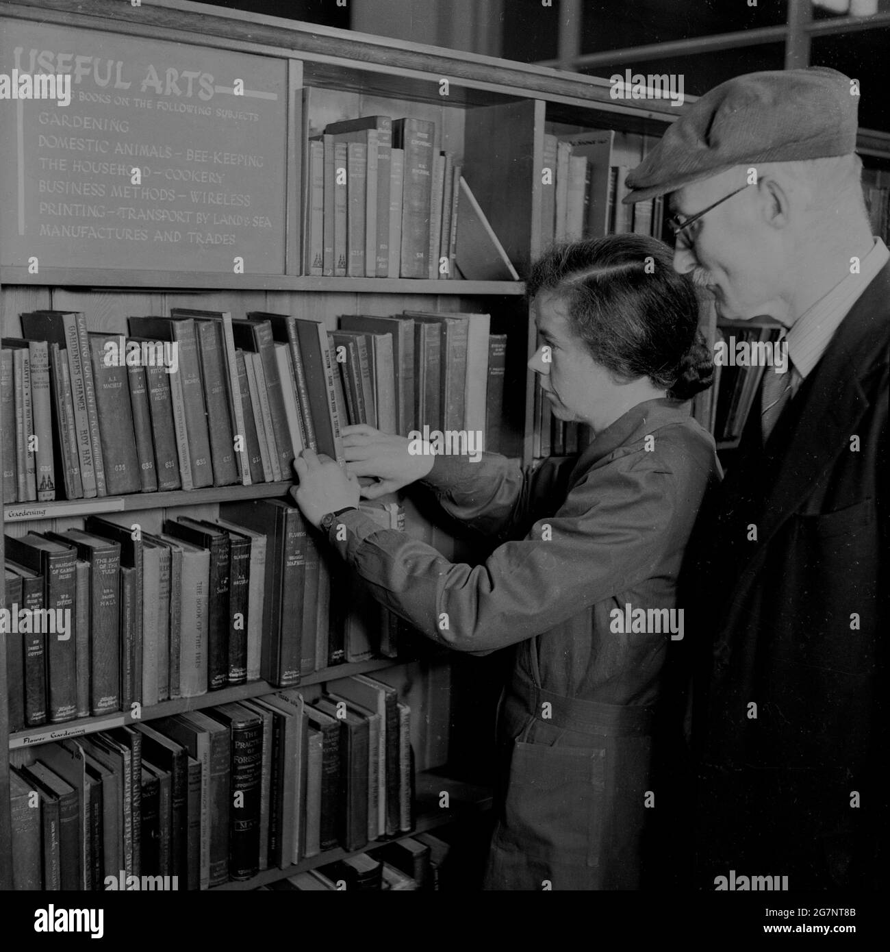 Anni '50, storico, all'interno di una biblioteca, una libaria femminile che mostra un anziano gentiluomo i libri disponibili nella sezione, 'Arti utili', Inghilterra, Regno Unito. Foto Stock
