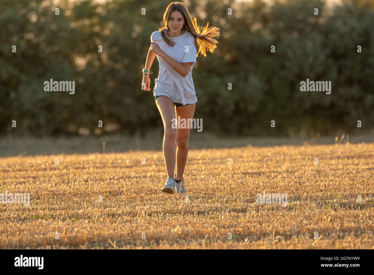 ragazza adolescente con lunghi capelli biondi, pantaloncini corti e t-shirt bianca che corre di fronte alla fotocamera attraverso un campo raccolto. Giocare sport sul campo. Foto Stock